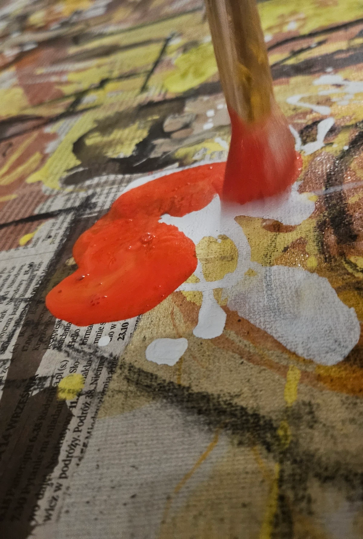 A brush applying a red stroke over newspaper collage and coloured layers on canvas.